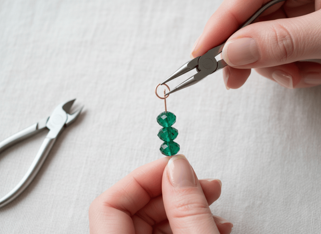 Hands using round-nose pliers to form a loop above an emerald bead stack.