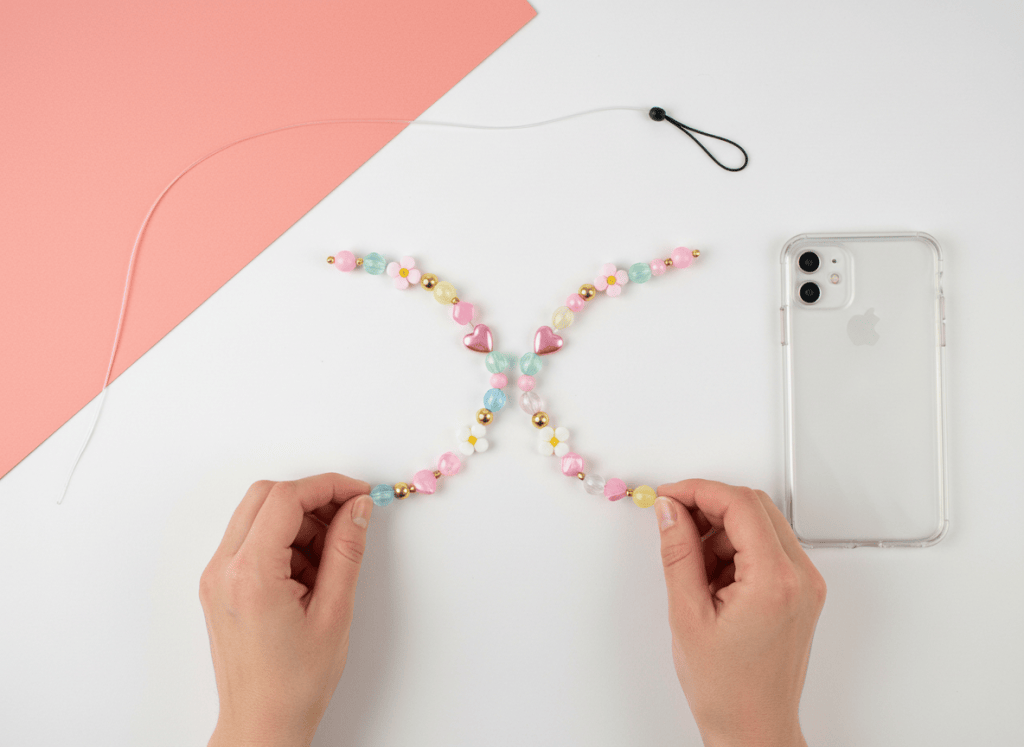 Hands arranging colorful translucent beads, charms, cord, and a phone charm loop beside a phone case.
